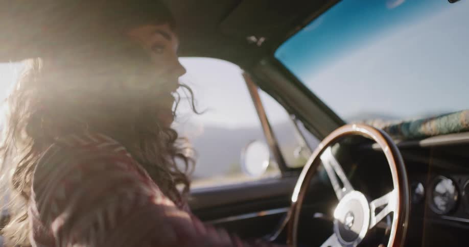 Sunlight flares as a joyful woman drives a classic car on a road trip, turning to high-five her friend