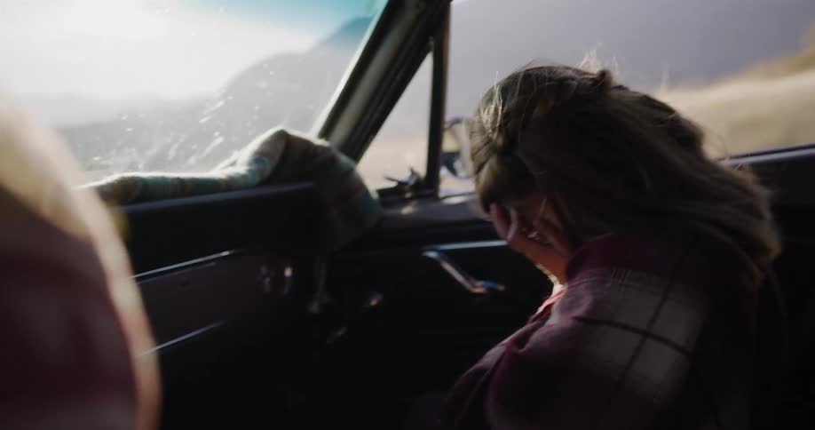 A joyful young girl wearing retro sunglasses laughs and smiles in the passenger seat of a classic convertible during a sunny road trip