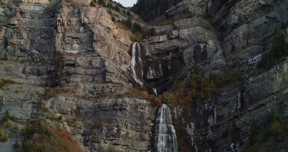 An aerial view reveals a magnificent waterfall cascading down a steep, rocky mountain cliff dotted with autumn-colored foliage