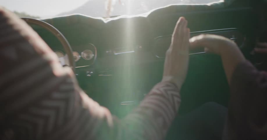 A carefree young couple in a vintage car play a hand-clapping game while on a sunny road trip through the mountains