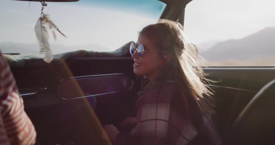 A smiling young woman with sunglasses enjoys a sunny road trip from the passenger seat of a classic convertible car