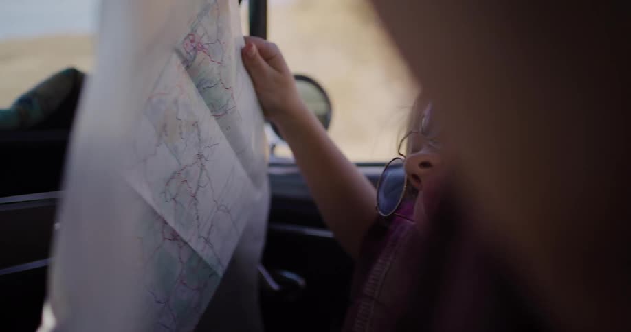 A happy young girl in blue sunglasses smiles as she reads a paper map in the passenger seat of a classic car on a desert road trip