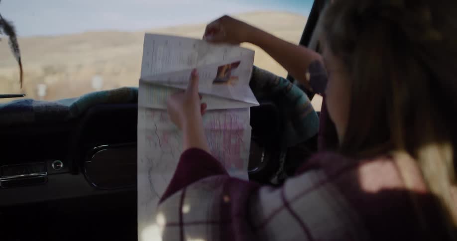 A young girl in the passenger seat of a classic car unfolds a paper road map for a road trip adventure in Utah