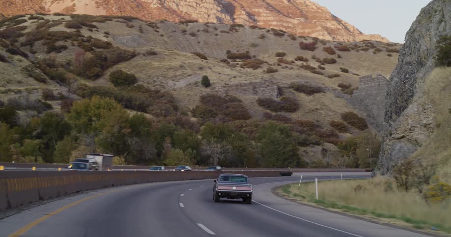 Rear view of a classic black muscle car driving on a scenic highway through a canyon at sunset