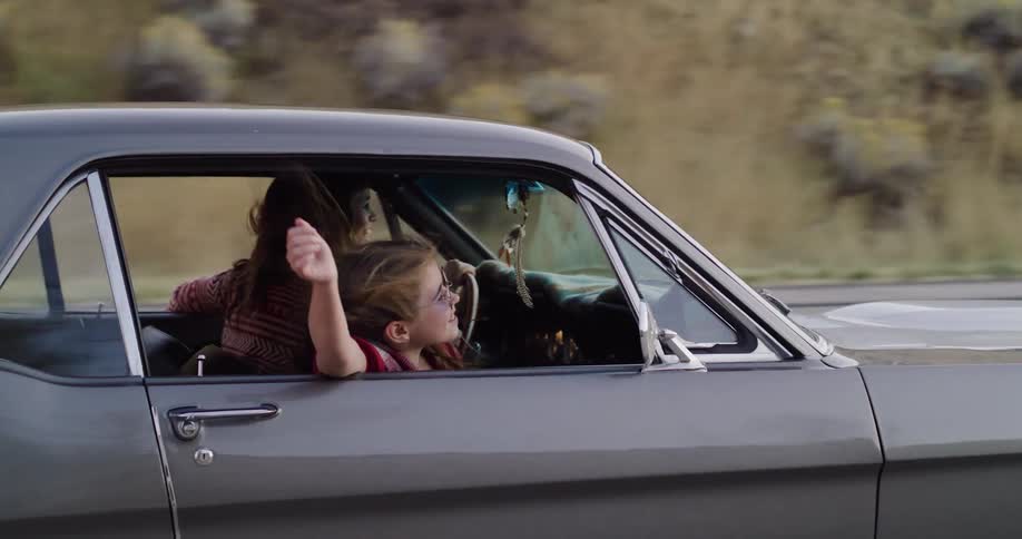 A happy young girl with sunglasses on sticks her head and hands out the window of a classic car as her mother drives down a country road at sunset