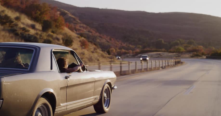 A carefree young woman drives a classic Mustang down a winding mountain road at sunset, waving her hand out the window in slow motion