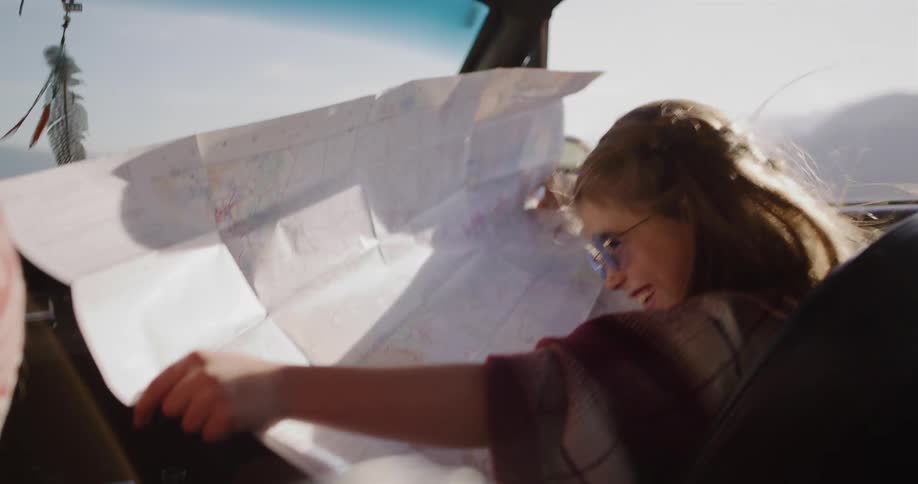 An excited young girl with sunglasses smiles as she navigates with a paper map during a sunny road trip in a vintage car