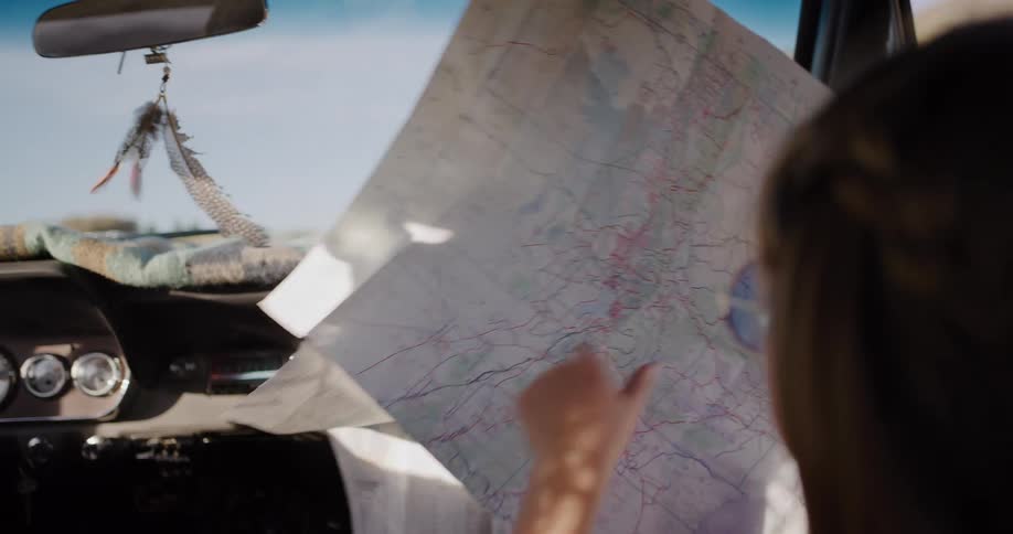 A young woman uses a magnifying glass to trace her route on a paper map while riding in a vintage car on a road trip