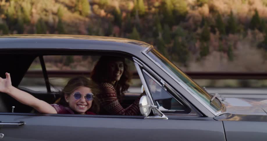 A carefree young girl with her arm out the window enjoys a scenic road trip with her mother in a vintage car