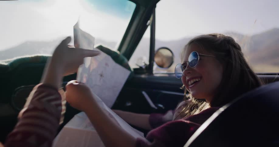 A smiling girl in sunglasses happily reads a paper map in the passenger seat of a vintage car during a sunny road trip