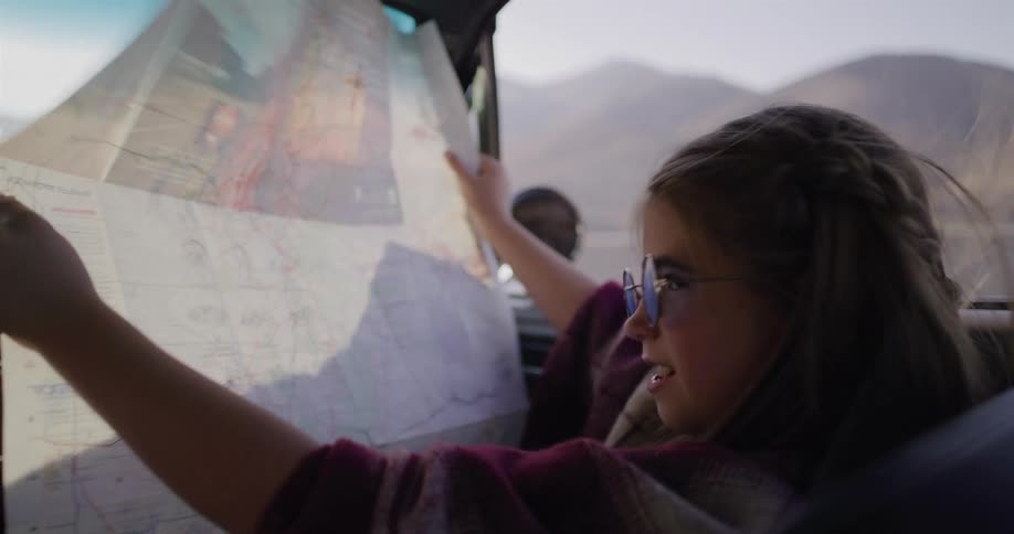A carefree girl wearing stylish sunglasses and a poncho smiles while navigating with a paper road map in a moving car during a desert road trip