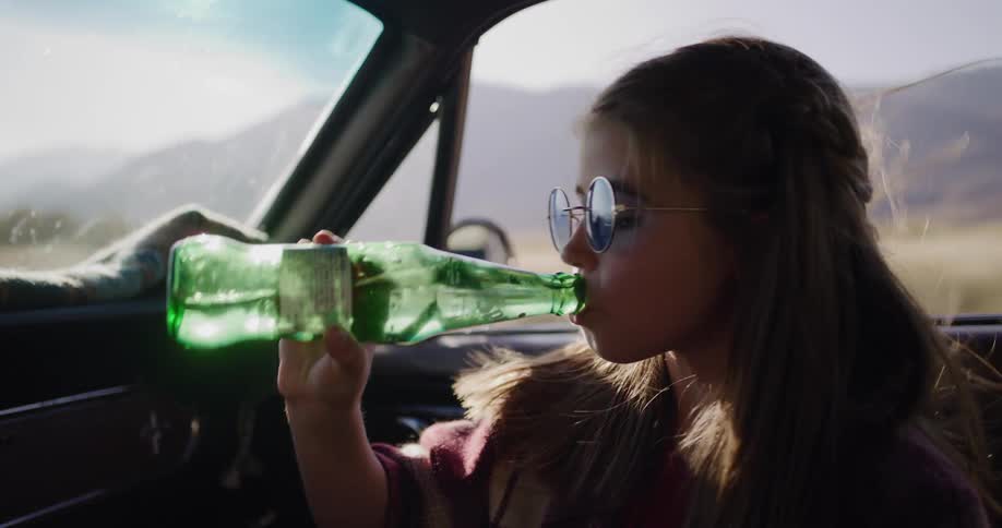 A cool young girl with blue sunglasses drinks from a glass bottle while on a sunny road trip through the desert
