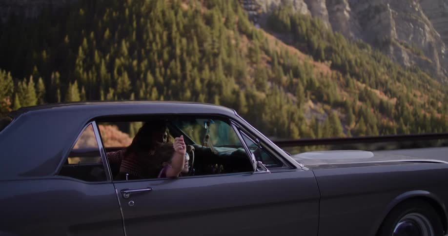 A happy young girl with sunglasses waves her hand out the window of a classic muscle car during a scenic mountain road trip