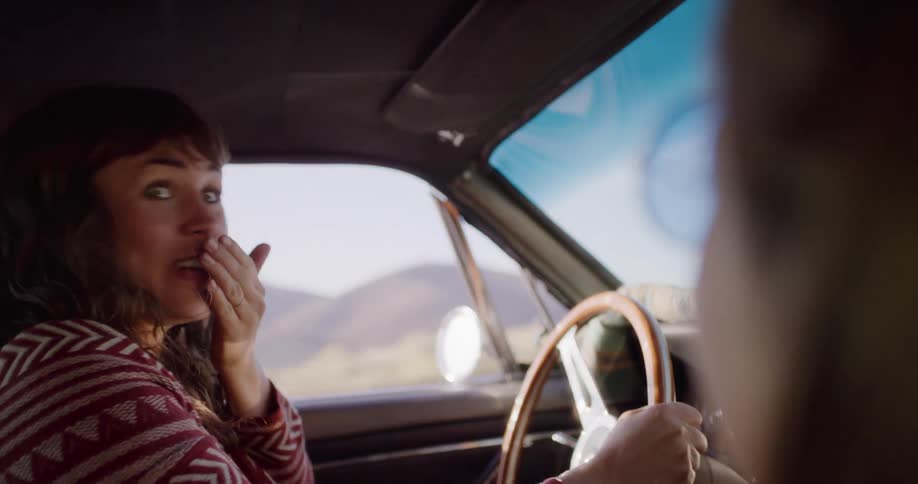 An expressive woman driving a vintage car on a road trip sings loudly, then laughs at her friend in the passenger seat