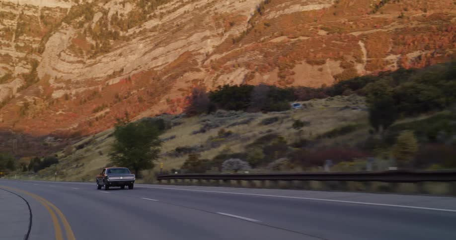 A vintage car cruises down a winding highway through a mountain canyon illuminated by the warm light of sunset