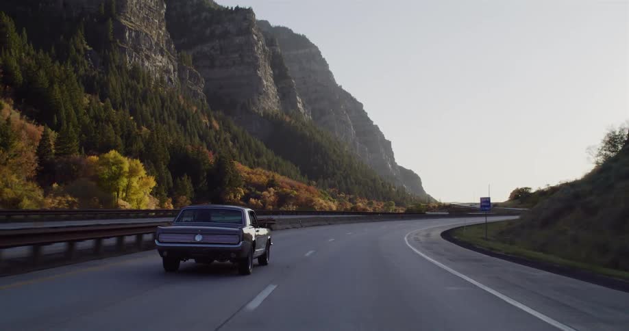 A classic muscle car cruises in slow motion down a scenic highway through a beautiful mountain canyon at sunrise