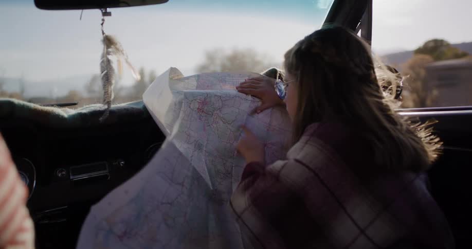 A young girl navigates using a paper map from the passenger seat of a vintage car during a sunny road trip through the countryside
