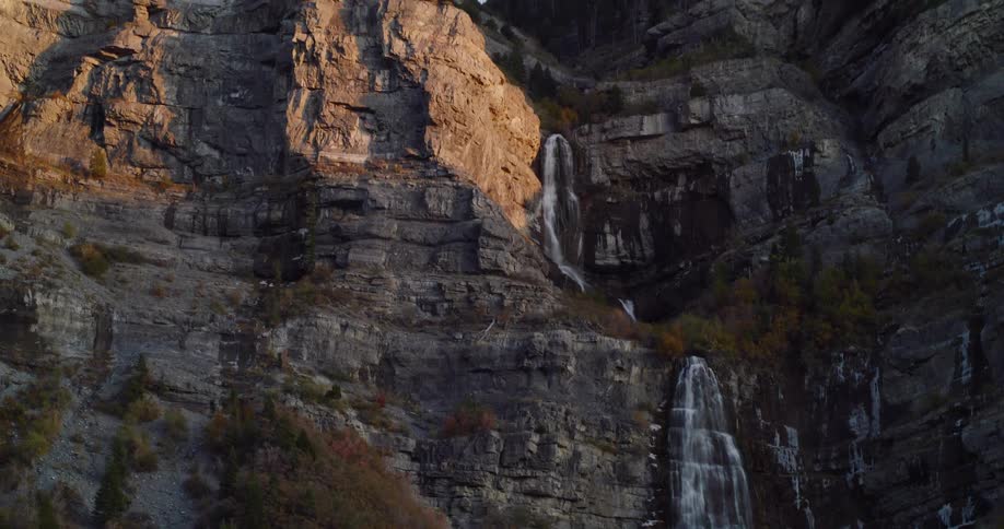 Upward pan of a beautiful multi-tiered waterfall on a rocky mountainside during a scenic drive at golden hour