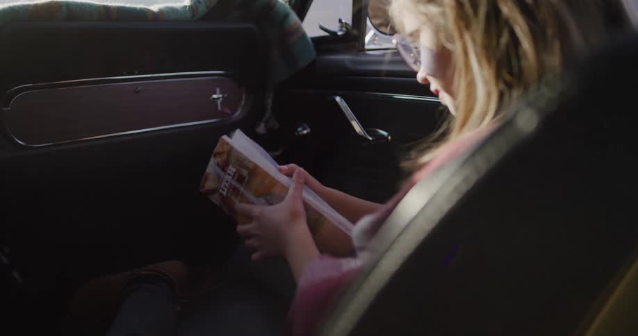 A young girl with glasses sits in the passenger seat of a classic car and eagerly unfolds a Utah road map to plan their adventure