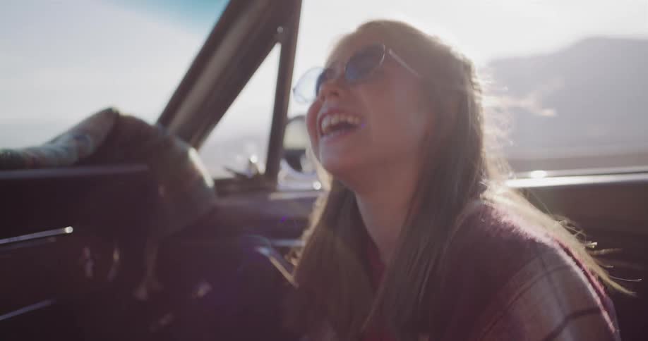 A young girl with round sunglasses smiles, laughs, and yawns in the passenger seat of a vintage car during a sunny road trip