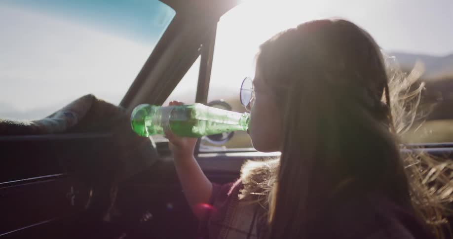A stylish young woman with retro sunglasses drinks a soda from a green bottle while riding in a classic convertible on a sunny day
