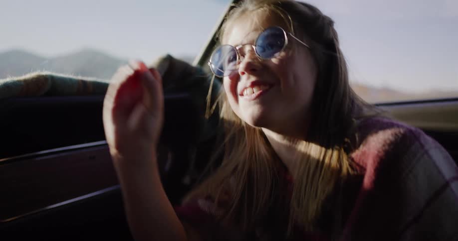 A joyful young girl in retro blue sunglasses laughs and eats candy while riding in a car on a sunny road trip