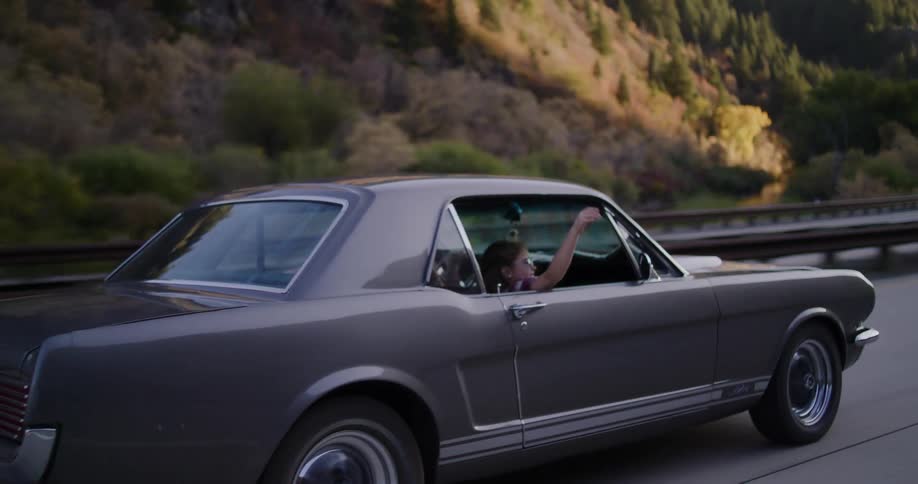 A carefree young woman waves her hand out the window of a classic grey Mustang while enjoying a scenic drive through the mountains