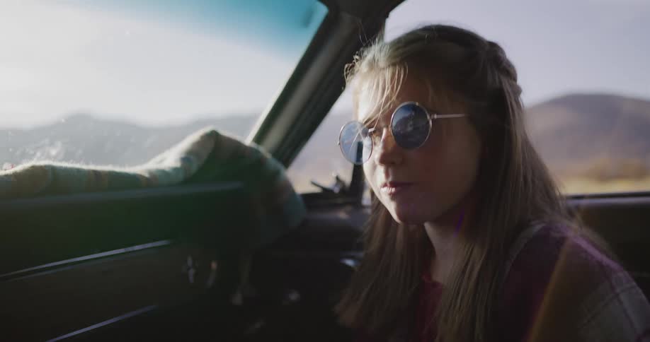 A carefree young girl in round sunglasses laughs with joy in slow motion while riding in a vintage car on a sunny road trip