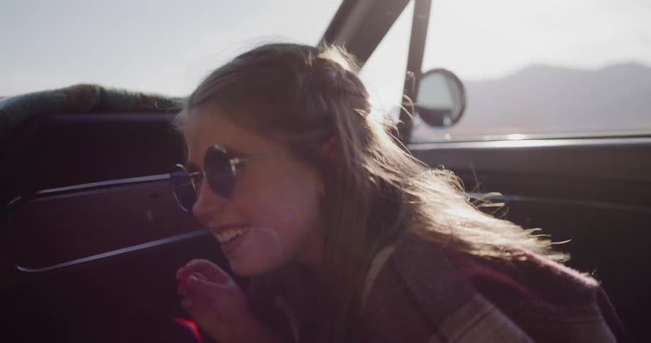 A free-spirited girl with round sunglasses laughs happily while enjoying a sunny road trip in a vintage car