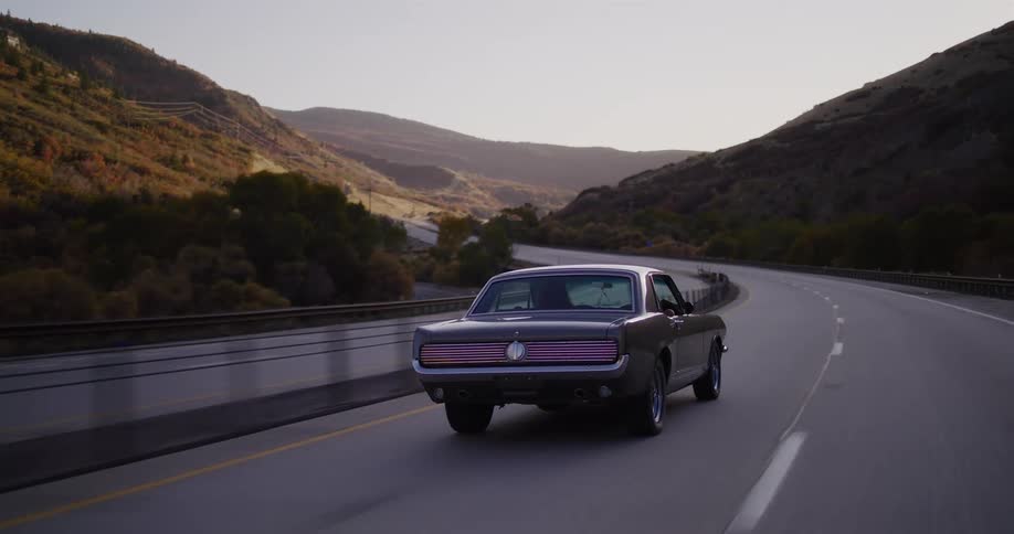 Rear view of a classic Ford Mustang driving along a winding highway through an autumn mountain landscape at dusk