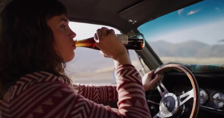 A woman drinks from a glass bottle of soda while driving a vintage car on a sunny day