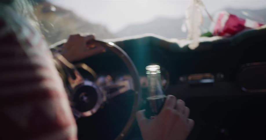 A woman's hand opens a glass soda bottle as she drives a classic car with the sun flaring through the windshield