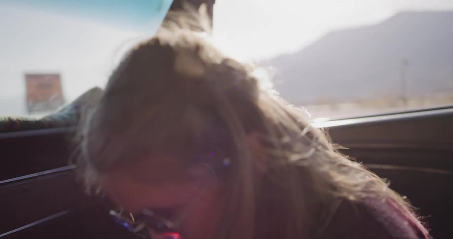 A happy young girl with blue sunglasses laughs and enjoys the ride from the passenger seat of a vintage car on a sunny road trip