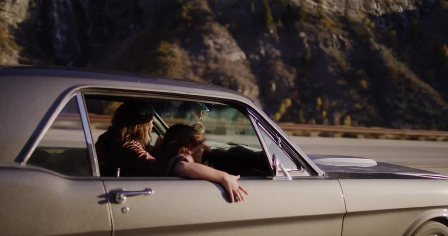 A free-spirited woman with sunglasses leans out of a classic car window, enjoying the wind as her friend drives along a scenic mountain highway