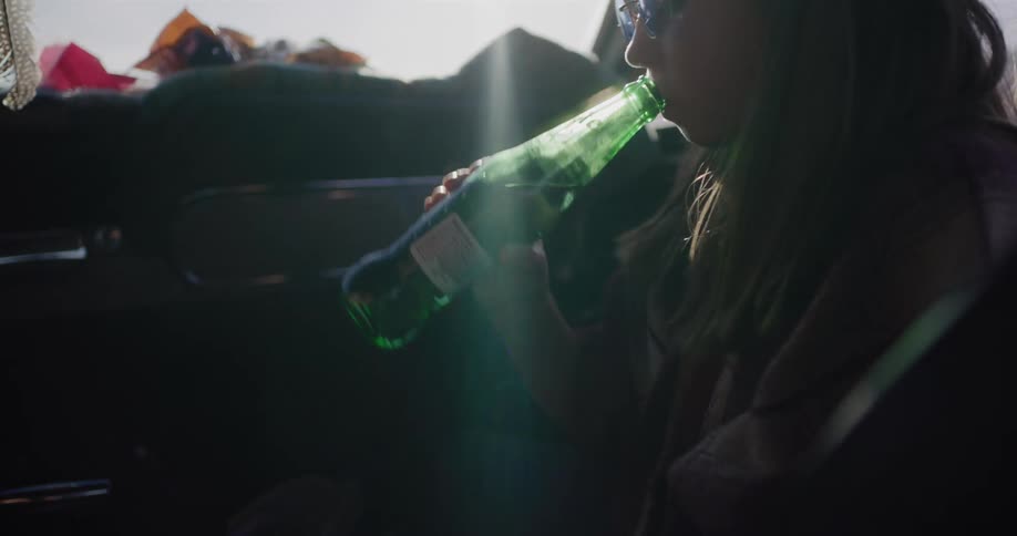 A woman takes a drink from a glass soda bottle while riding in the passenger seat of a vintage car on a sunny day