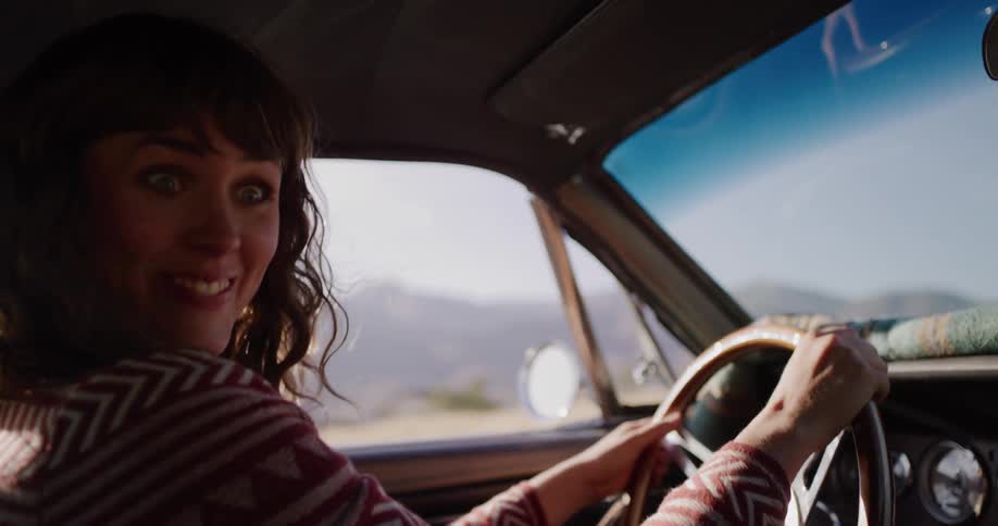 An expressive woman sings, yells, and then gives a playful look to her passenger while driving a vintage car on a sunny road trip