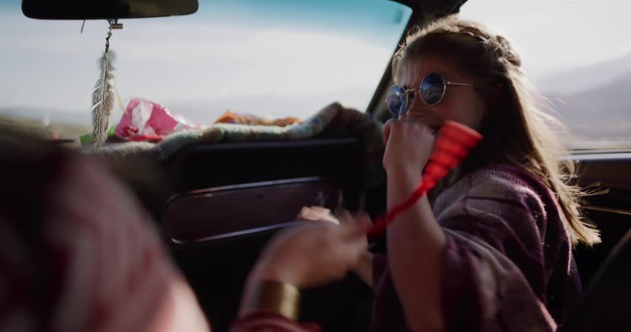 A carefree girl in a vintage car playfully shares a red licorice rope with her mom on a sunny road trip