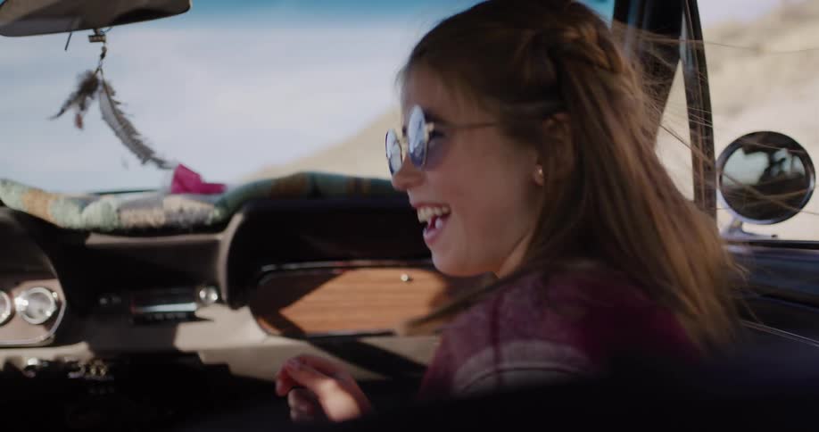 A joyful young girl laughs and eats a raspberry in the passenger seat of a vintage car during a sunny desert road trip