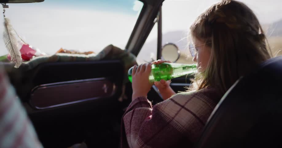 Young girl in sunglasses drinks a soda in the passenger seat of a vintage car during a sunny road trip