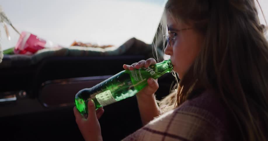 A man and a young girl toast with glass soda bottles inside a vintage car on a sunny road trip