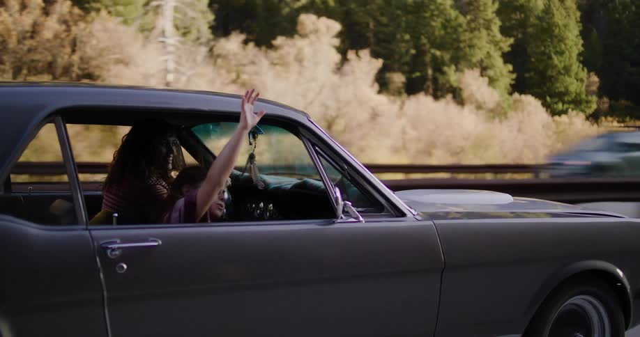 A joyful young girl with sunglasses feels the freedom of the open road, waving her hand out the window of a classic grey Mustang