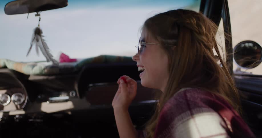 A slow-motion close-up captures a joyful young woman in sunglasses laughing as she rides in the passenger seat of a vintage car on a sunny day