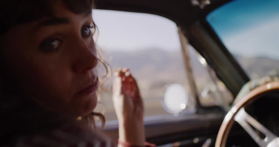 A woman drives a classic car through the sunny countryside while smoking a hand-rolled cigarette