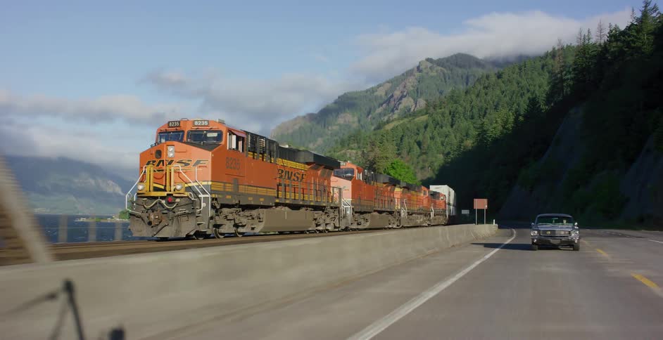 Driving shot tracking a BNSF freight train as it moves parallel to a classic car on a scenic highway next to a river and mountains