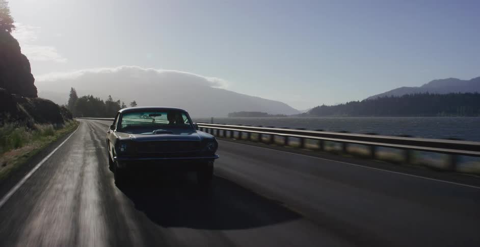 A classic black muscle car cruises down a scenic coastal highway at sunrise