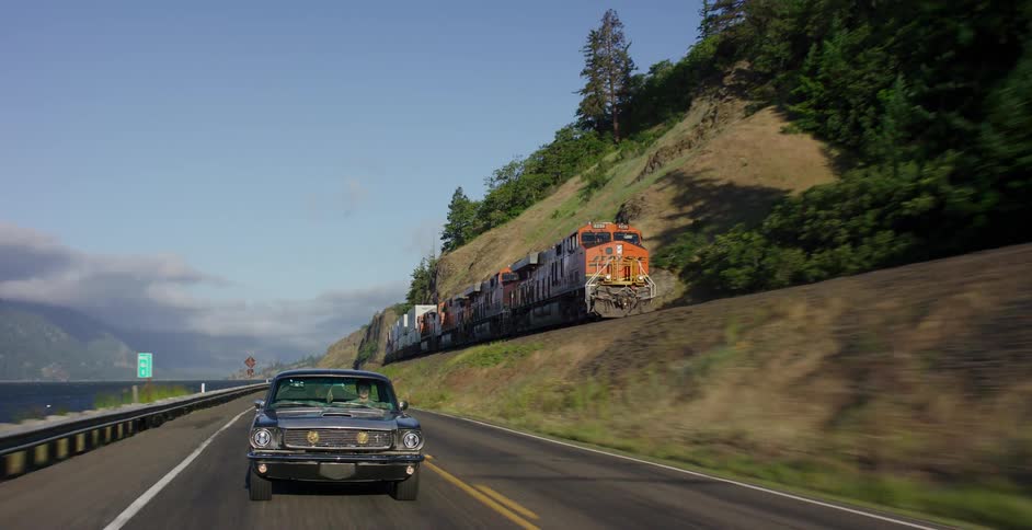 A classic muscle car drives along a scenic highway next to a river, with a long freight train moving on parallel tracks up on the hillside