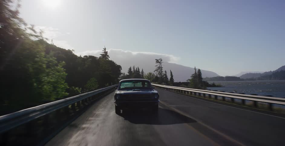 A classic black muscle car cruises down a scenic highway alongside a vast river with mountains in the distance on a sunny day