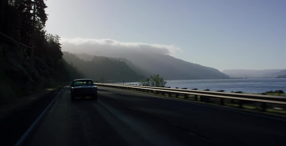 A classic black muscle car cruises down a scenic highway alongside a misty river gorge at sunrise