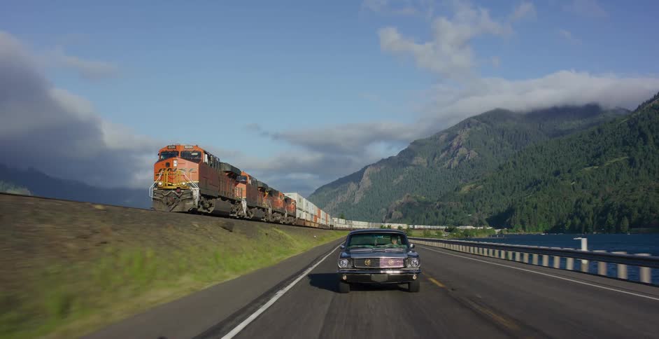 A classic convertible cruises down a scenic highway next to a river, racing alongside a massive freight train with mountains in the background