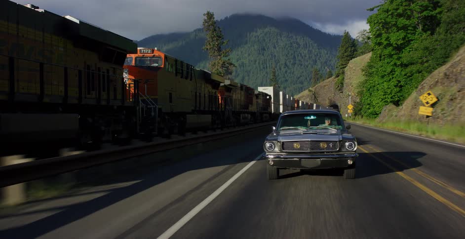 A classic muscle car drives along a scenic mountain road next to a moving freight train